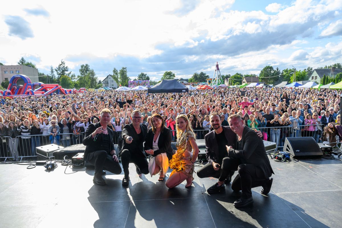 Zespół na scenie, z tłumem w tle; niebo błękitne, scenografia kolorowa. Tłoczna publika, atmosfera festiwalu.