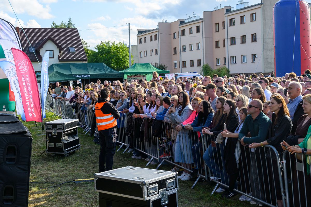 Tłum zbiera się przed sceną. Osoba w kamizelce odblaskowej stoi z przodu. W tle widać budynki i namioty.
