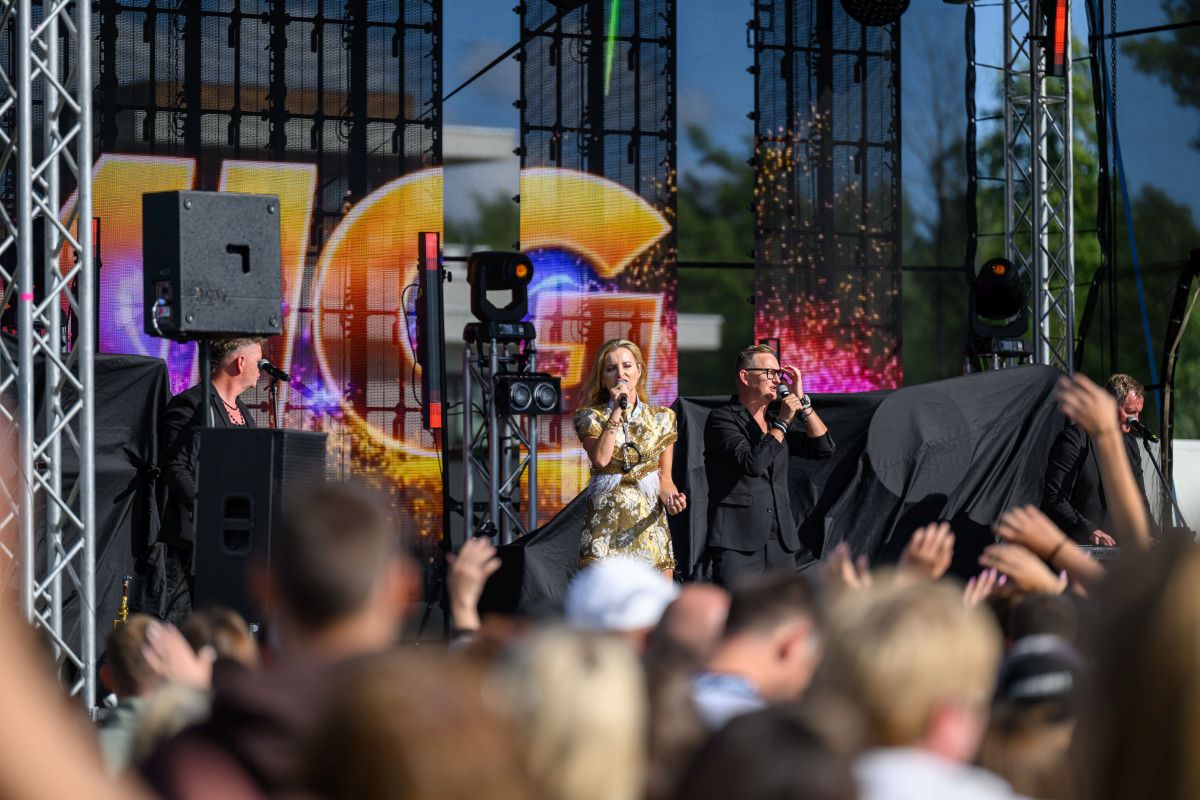 Koncert na scenie plenerowej, kobieta i mężczyzna śpiewają, tło z kolorowymi efektami świetlnymi. Publikum klaszcze, dzień słoneczny.