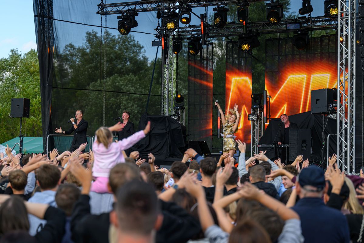 Koncert na zewnątrz, tłum wspólnie się bawi. Scena z artystami w czerni i kolorowym świetle. Zielone drzewa w tle.