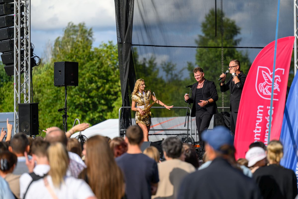 Scena plenerowa z występem muzycznym, kobieta w złotej sukience trzyma saksofon. Tło zielone drzewa, nieboj znak. Ludzie w tłumie.