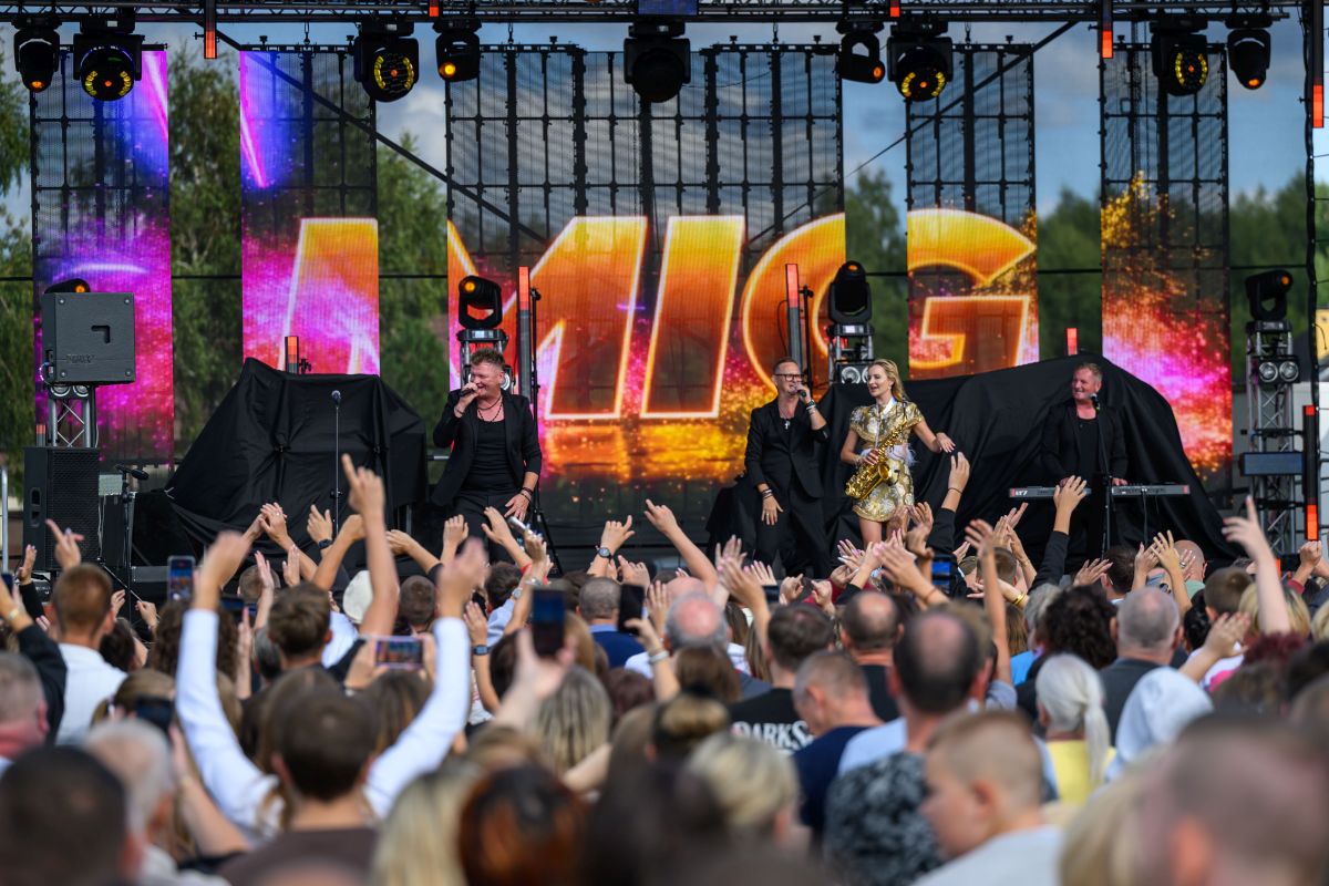 Koncert na zewnątrz z tłumem ludzi pod sceną. Jasne, kolorowe światła i ekran z napisem w tle. Cztery osoby na scenie: trzech mężczyzn i jedna kobieta.