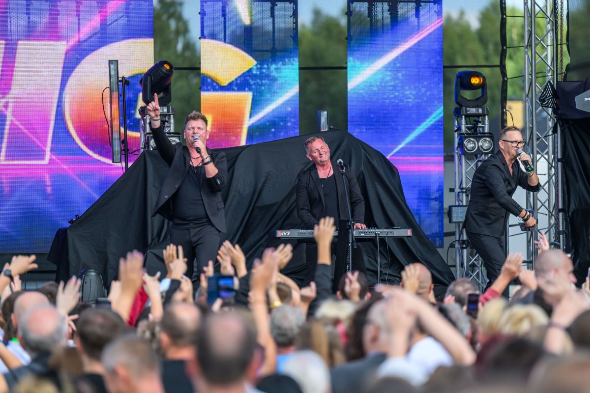 Trzech muzyków w czarnych strojach występuje na scenie. Tło to kolorowe ekrany LED z abstrakcyjnymi wzorami. Publiczność podnosi ręce w rytm muzyki.