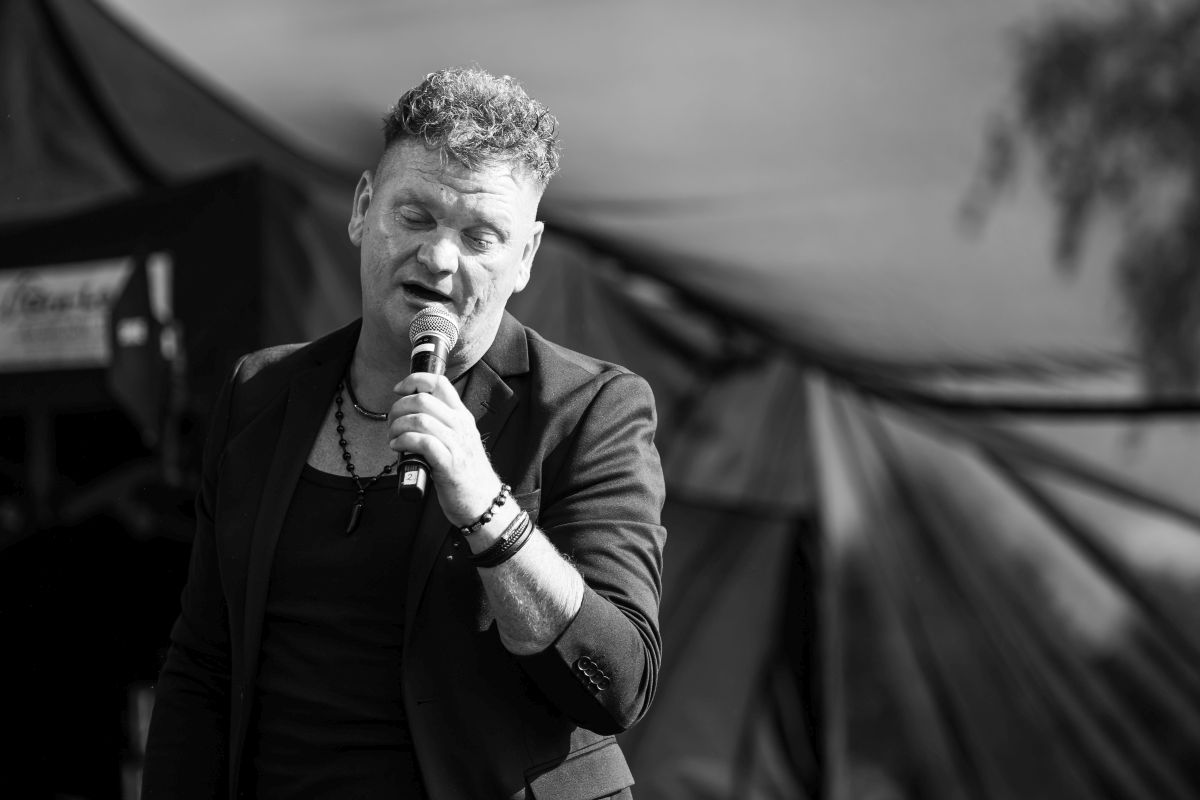 Czarno-białe zdjęcie przedstawia mężczyznę śpiewającego do mikrofonu, w tle rozmyte namioty. Ma krótkie kręcone włosy i nosi ciemne ubranie.