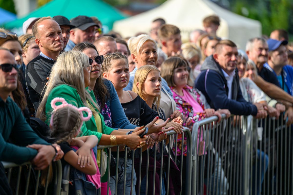 Na zdjęciu widać grupę ludzi za barierkami, spoglądających przed siebie. Ubrani w różnorodne kolory, na tle zieleni i namiotów. Atmosfera przypomina festyn lub wydarzenie na świeżym powietrzu.