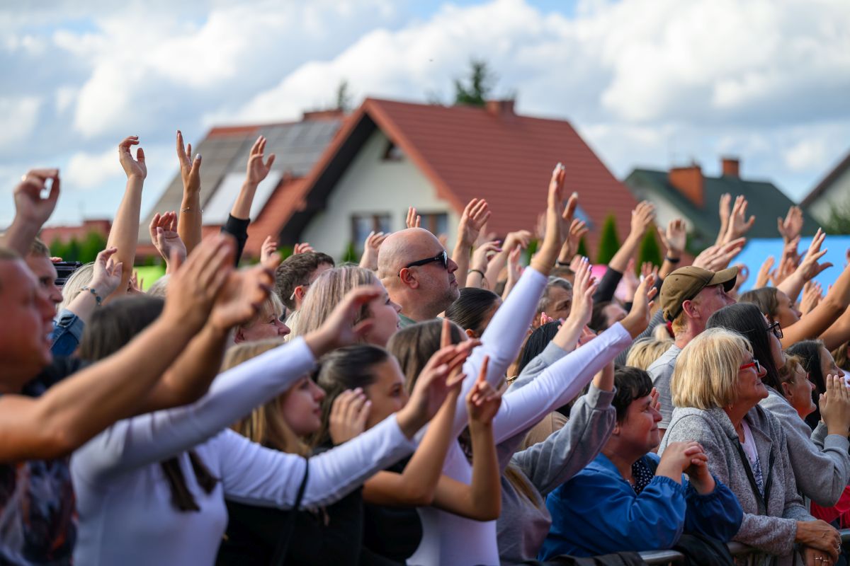 Tłum rozradowanych ludzi z podniesionymi rękoma na tle domów z czerwonymi dachami i nieba z chmurami.