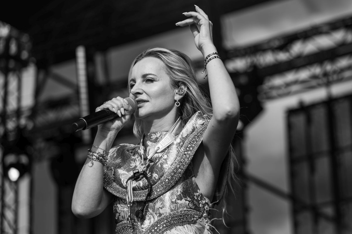Kobieta na scenie śpiewa, trzymając mikrofon. Jest w ozdobnym stroju, z podniesioną ręką. Za nią rozmyte tło sceny.