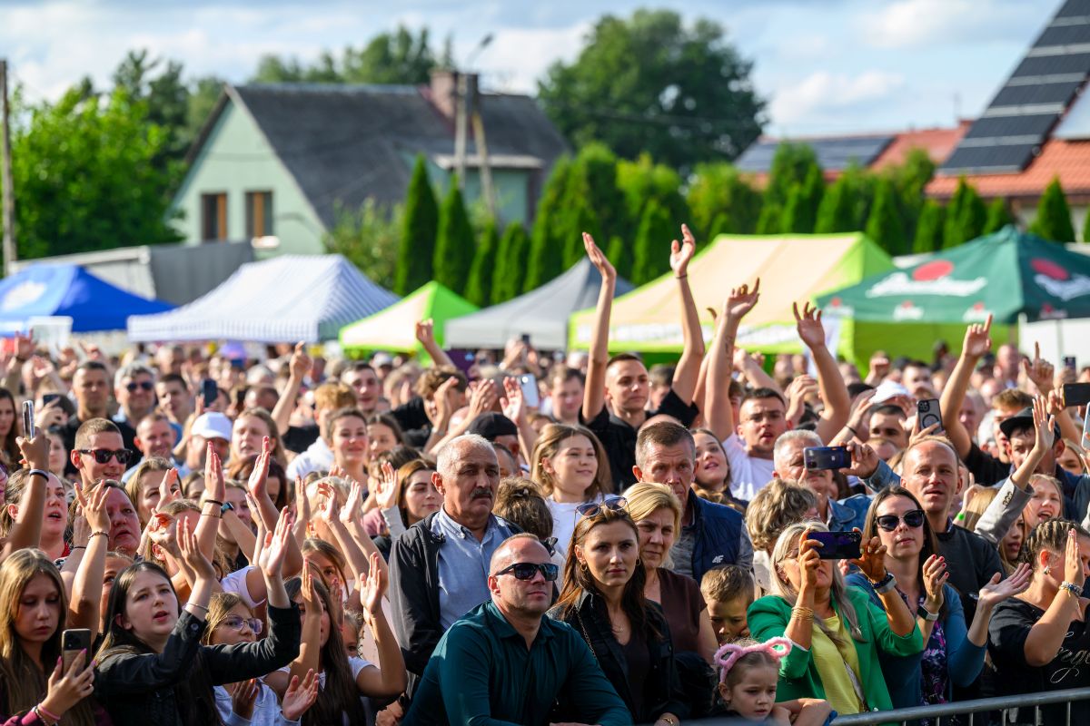 Na zdjęciu tłum ludzi na zewnątrz, uniesione ręce. Kolorowe namioty w tle, zielona roślinność i kilka budynków. Dzień jest słoneczny.