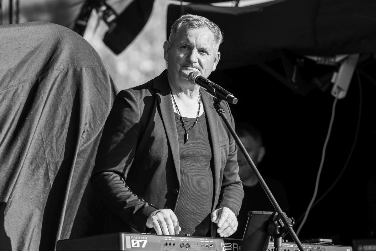 Na czarno-białym zdjęciu mężczyzna przy mikrofonie gra na klawiszach. Ubrany w ciemną marynarkę i naszyjnik. W tle ciemne zasłony sceniczne.