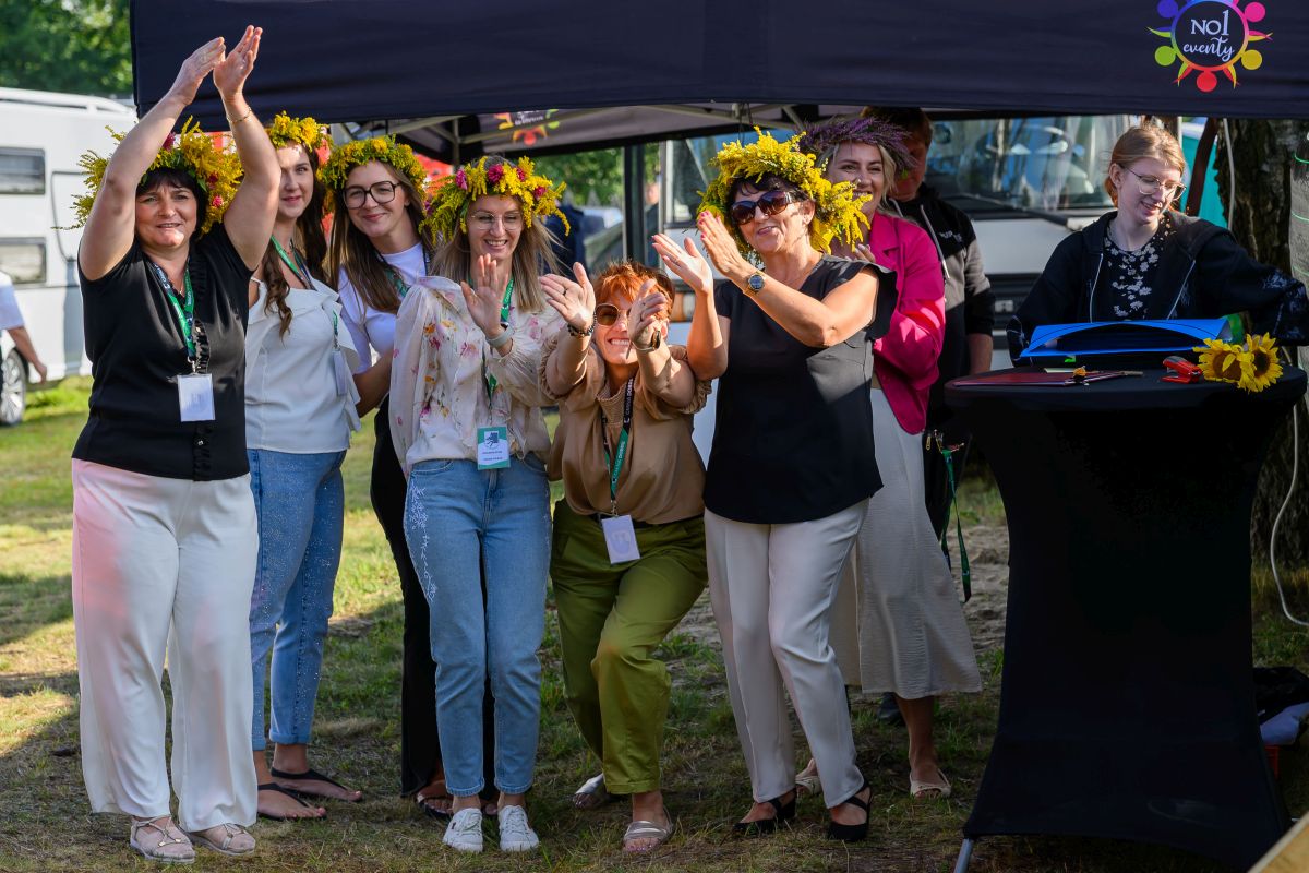 Grupa osób w wiankach z kwiatów uśmiecha się i klaska pod czarnym namiotem. Noszą kolorowe ubrania, tło jest zielone. Atmosfera jest radosna.