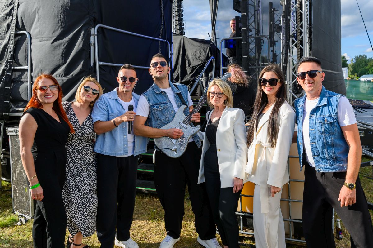 Grupa osób stoi na zewnątrz przy scenie, ubrana w kolorowe stroje, w tym jeans i biel. Na ziemi trawa, a za nimi czarne tło. Jedna osoba trzyma gitarę, a inna mikrofon.
