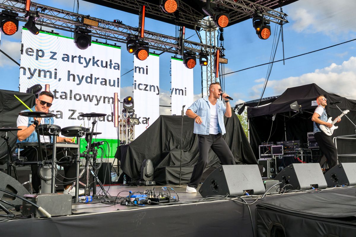 Na scenie trzech muzyków, w tle duże ekrany z tekstem i jasno-niebieskie niebo. Są światła sceniczne i sprzęt muzyczny. Artyści ubrani w jasne koszule i czarne spodnie.