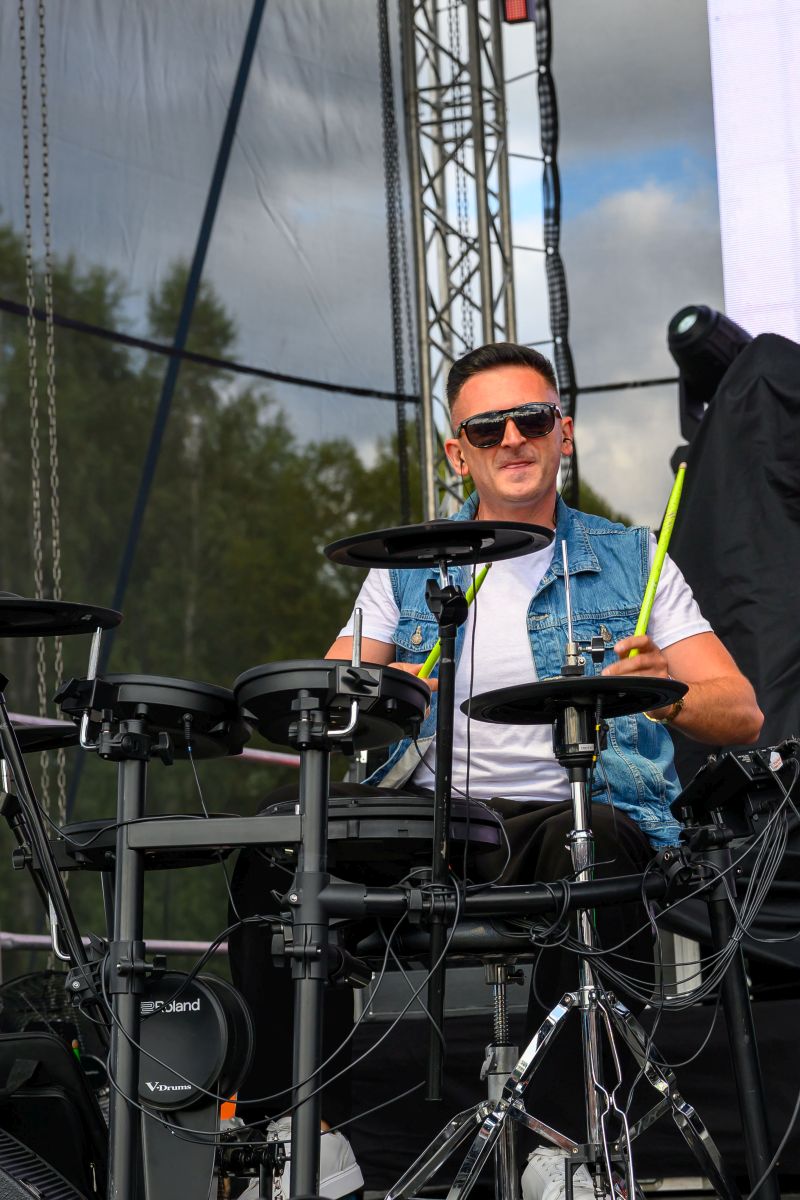 Mężczyzna gra na elektronicznej perkusji na scenie. Nosi jeansową kamizelkę i ciemne okulary. Tło tworzy niebo i sprzęt sceniczny.