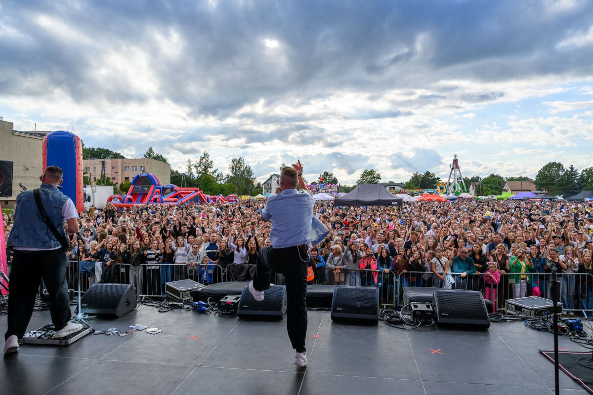 Na scenie muzycy występują przed tłumem. Niebo pochmurne, ale jasne, w tle widać kolorowe atrakcje plenerowe. Publiczność entuzjastycznie reaguje, tworząc dynamiczną atmosferę koncertu.