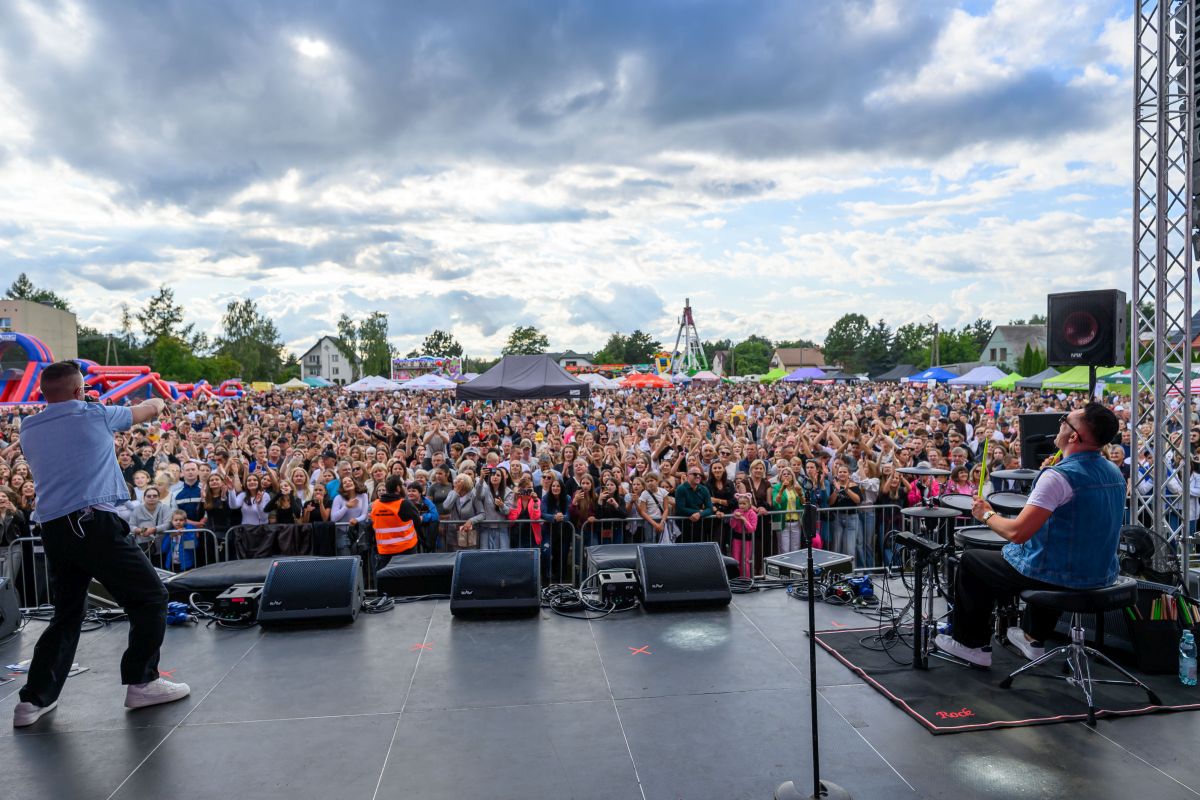 Koncert na świeżym powietrzu, z tłumem widzów pod sceną. Występujący artysta w jasnej koszuli, niebo zachmurzone. Na scenie sprzęt muzyczny, a w tle rozrywkowe budki.