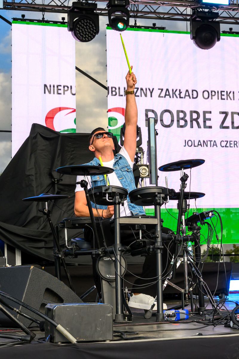 Mężczyzna w okularach przeciwsłonecznych gra na elektronicznej perkusji na scenie. W tle widoczne plakaty i reflektory. Kolory to głównie czerń, biel i zieleń.