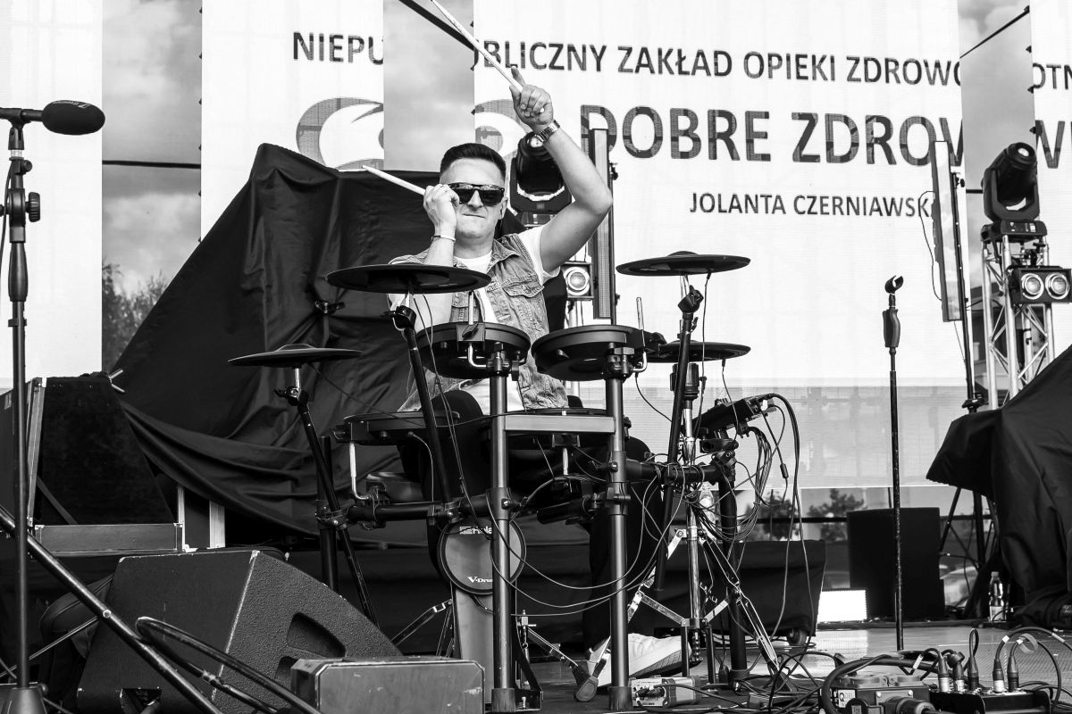 Mężczyzna gra na elektronicznej perkusji. Tło zawiera baner z napisami i sprzęt sceniczny. Zdjęcie jest czarno-białe.