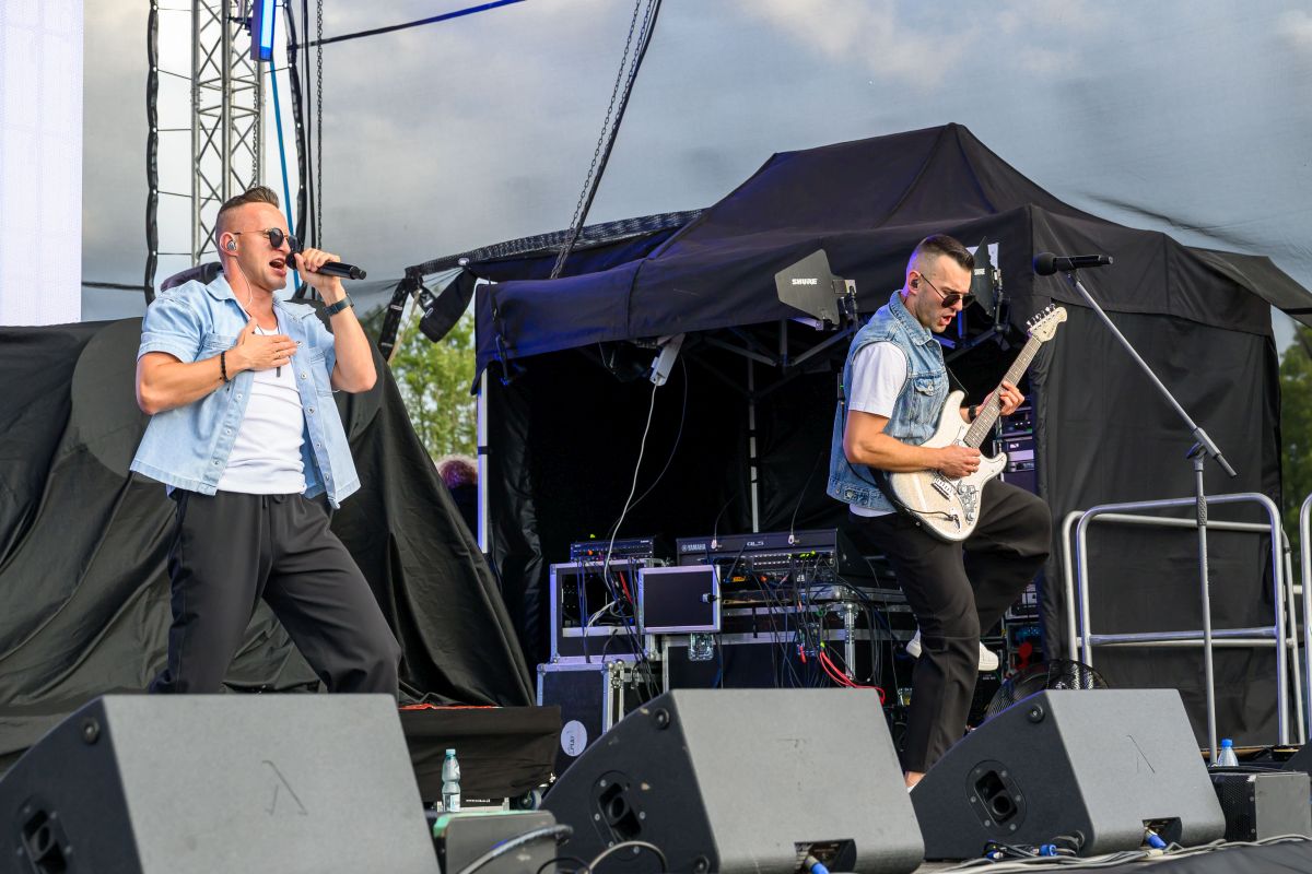 Dwóch mężczyzn występuje na scenie. Jeden śpiewa, drugi gra na gitarze elektrycznej. Tło jest ciemne z sprzętem scenicznym.