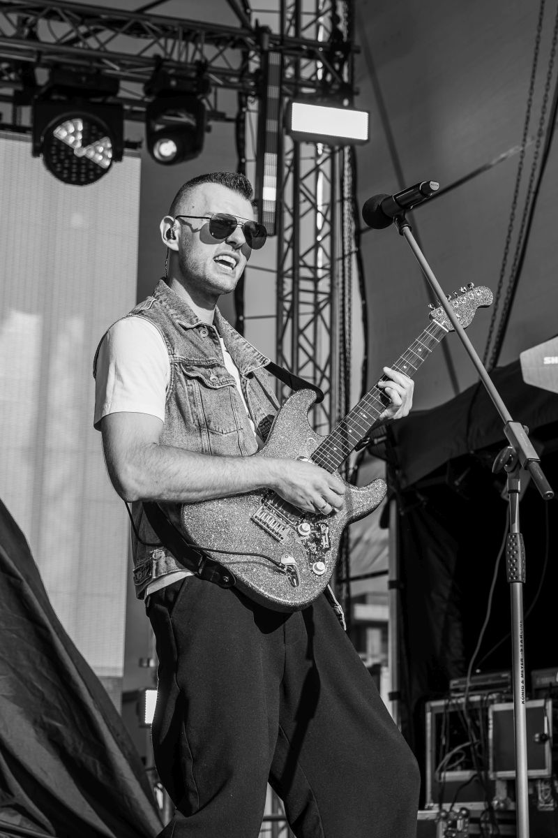 Mężczyzna gra na gitarze elektrycznej na scenie, uśmiecha się, nosi okulary przeciwsłoneczne i kamizelkę jeansową. Tło to scenografia koncertowa z oświetleniem. Zdjęcie jest czarno-białe.