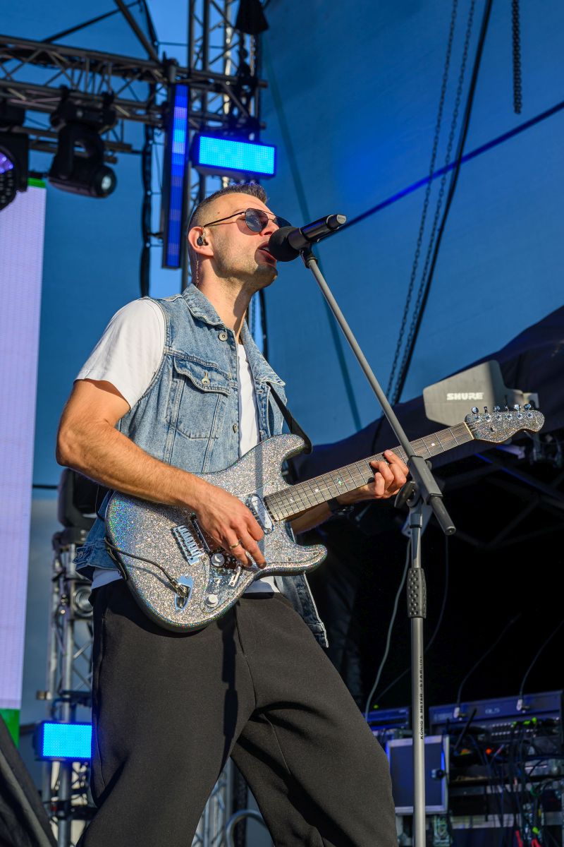 Zespół na scenie; muzyk w okularach śpiewa, trzymając połyskującą gitarę. Tło jest niebieskie, oświetlone reflektorami.