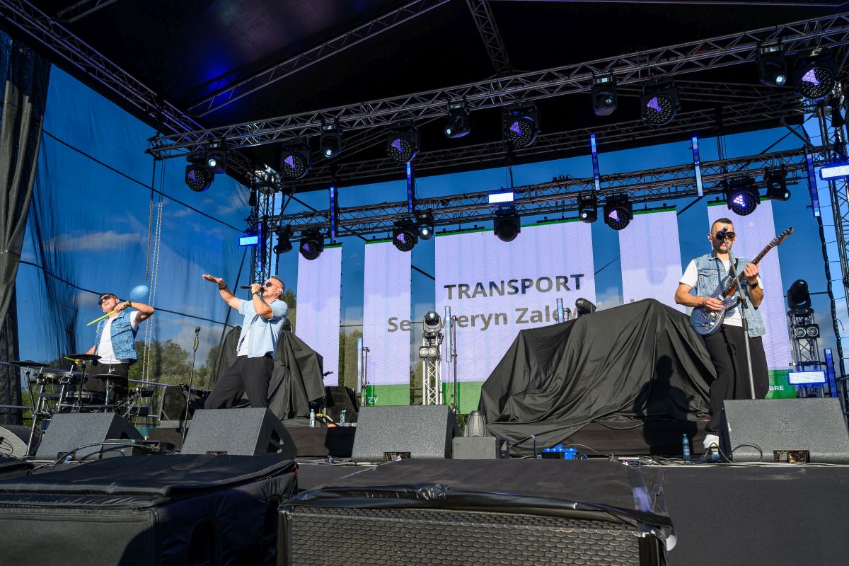 Trzech muzyków na scenie grających na instrumentach. Tło z niebieskim niebem i konstrukcją sceny. Napis TRANSPORT na ekranie.