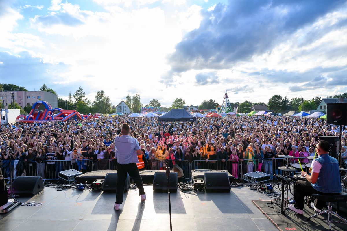 Koncert na świeżym powietrzu, widziany z perspektywy sceny. Liczna publiczność na tle nieba z chmurami. Kolorowe stoiska i zjeżdżalnia widoczne z tyłu.