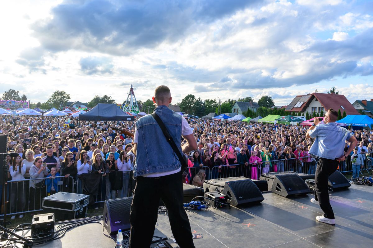 Na zdjęciu jest koncert na świeżym powietrzu z dwoma muzykami na scenie. Tłum ludzi i kolorowe stoiska w tle. Niebo jest pochmurne.