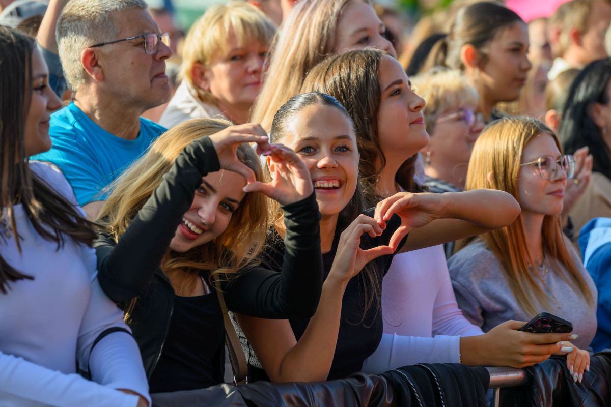Na zdjęciu grupa uśmiechniętych osób na koncercie, pokazujących serca dłońmi. Przeważają jasne i ciepłe kolory. W tle widać tłum ludzi.