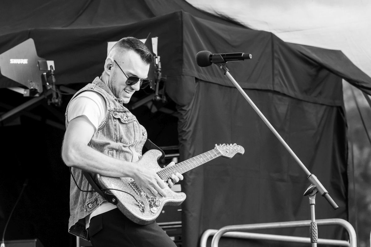 Mężczyzna gra na gitarze elektrycznej na scenie. Nosi okulary przeciwsłoneczne i dżinsową kamizelkę. Tło to ciemne kurtyny sceniczne.