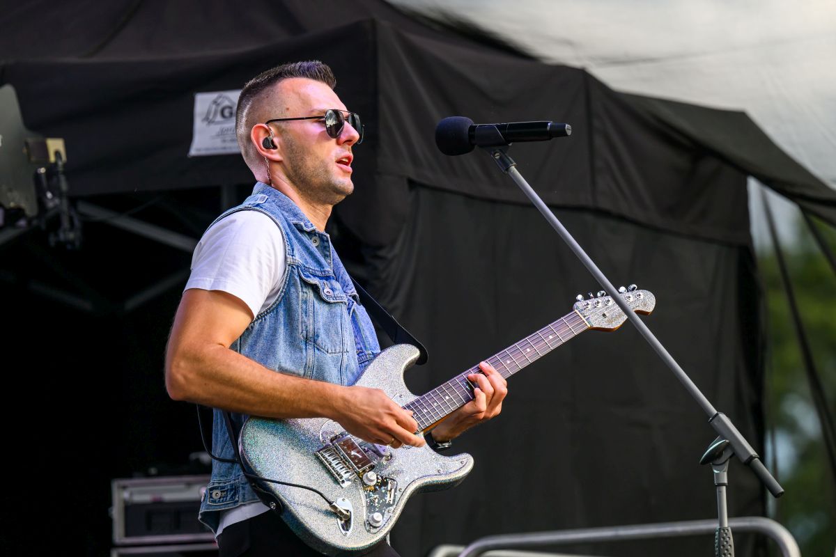 Mężczyzna gra na elektrycznej gitarze, nosi dżinsową kamizelkę. Tło sceny jest ciemne. Śpiewa do mikrofonu na statywie.