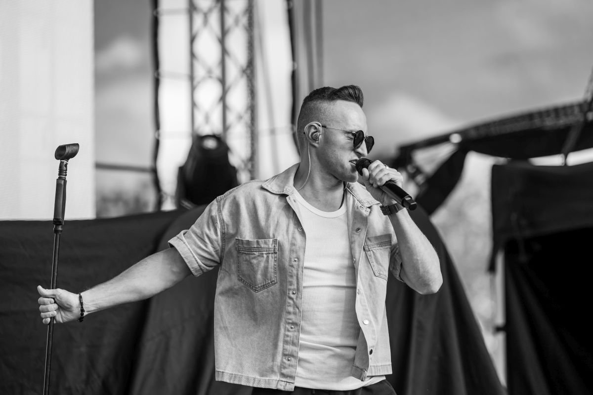 Mężczyzna w okularach przeciwsłonecznych śpiewa na scenie, trzyma mikrofon. Na sobie ma dżinsową koszulę. W tle rozmyte elementy sceny.