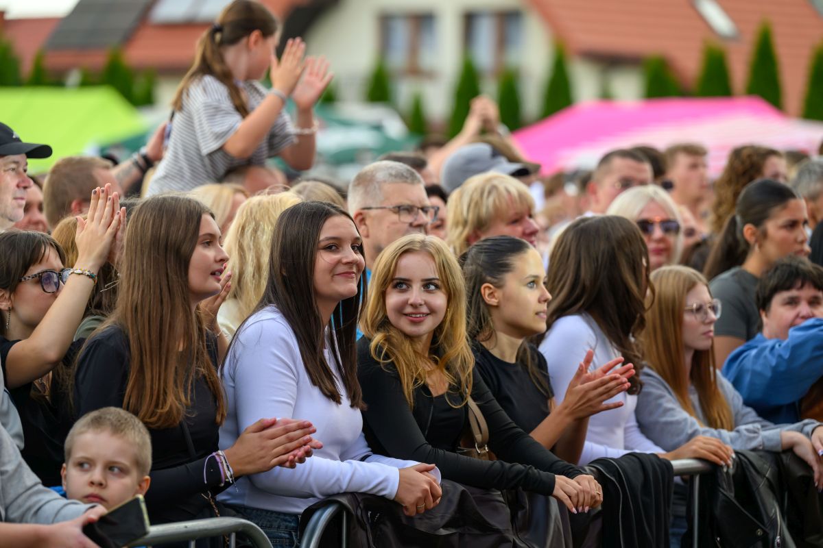 Grupa ludzi na imprezie na świeżym powietrzu, w jasnych i czarnych ubraniach. Tło to kolorowe namioty. Osoby wyglądają na zadowolone i klaszczą.