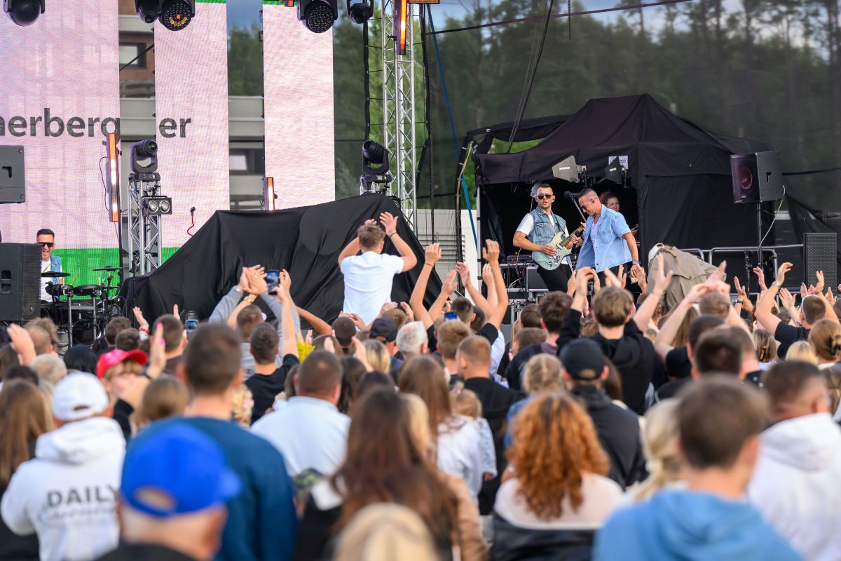 Na zdjęciu koncert na świeżym powietrzu, ludzie pod sceną podnoszą ręce. Na scenie muzycy z gitarą, z tyłu las. Dominuje kolor czarny i niebieski.