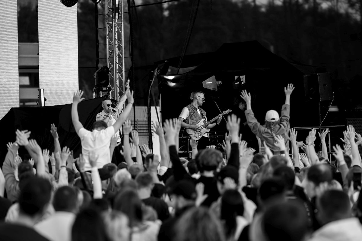 Na scenie muzycy grają koncert, otoczeni tłumem z uniesionymi rękoma. Tło jest ciemne, z widoczną konstrukcją sceny. Fotografia jest czarno-biała.