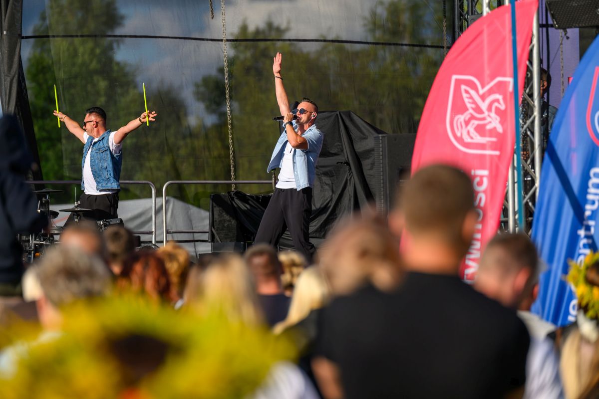 Na zdjęciu dwaj mężczyźni na scenie, jeden śpiewa. Obok nich kolorowe flagi, w tle zielone drzewa. Przed sceną widownia.