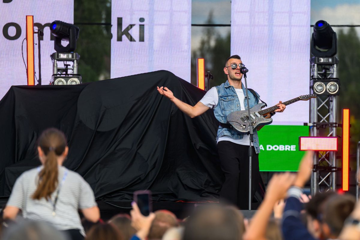 Mężczyzna z gitarą w okularach śpiewa na scenie. Ma na sobie jeansową kamizelkę, z tyłu kolorowe światła. Przed nim tłum ludzi.