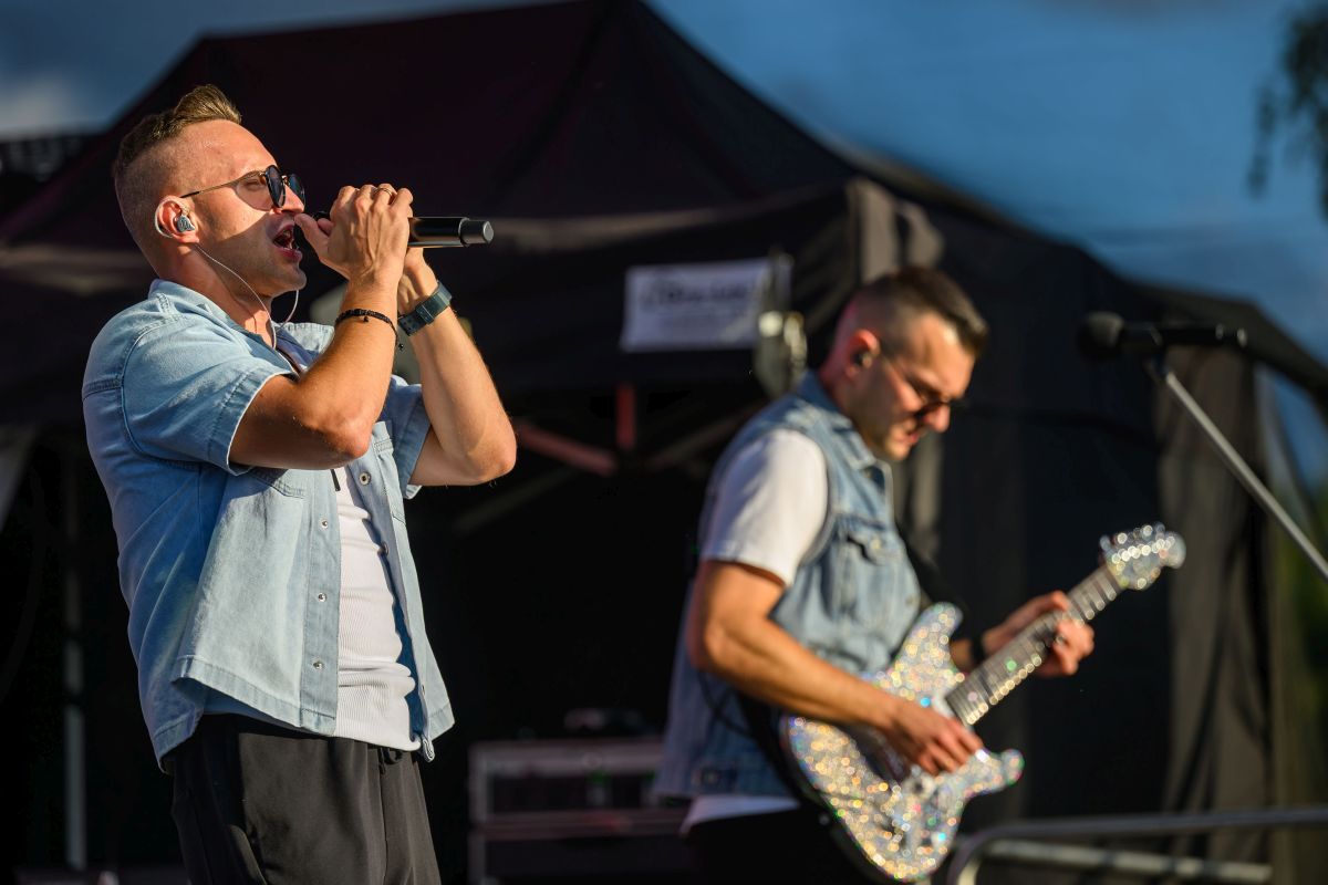 Dwóch mężczyzn na scenie. Jeden śpiewa do mikrofonu, drugi gra na błyszczącej gitarze. Tło jest ciemne, a ich ubrania jasne.