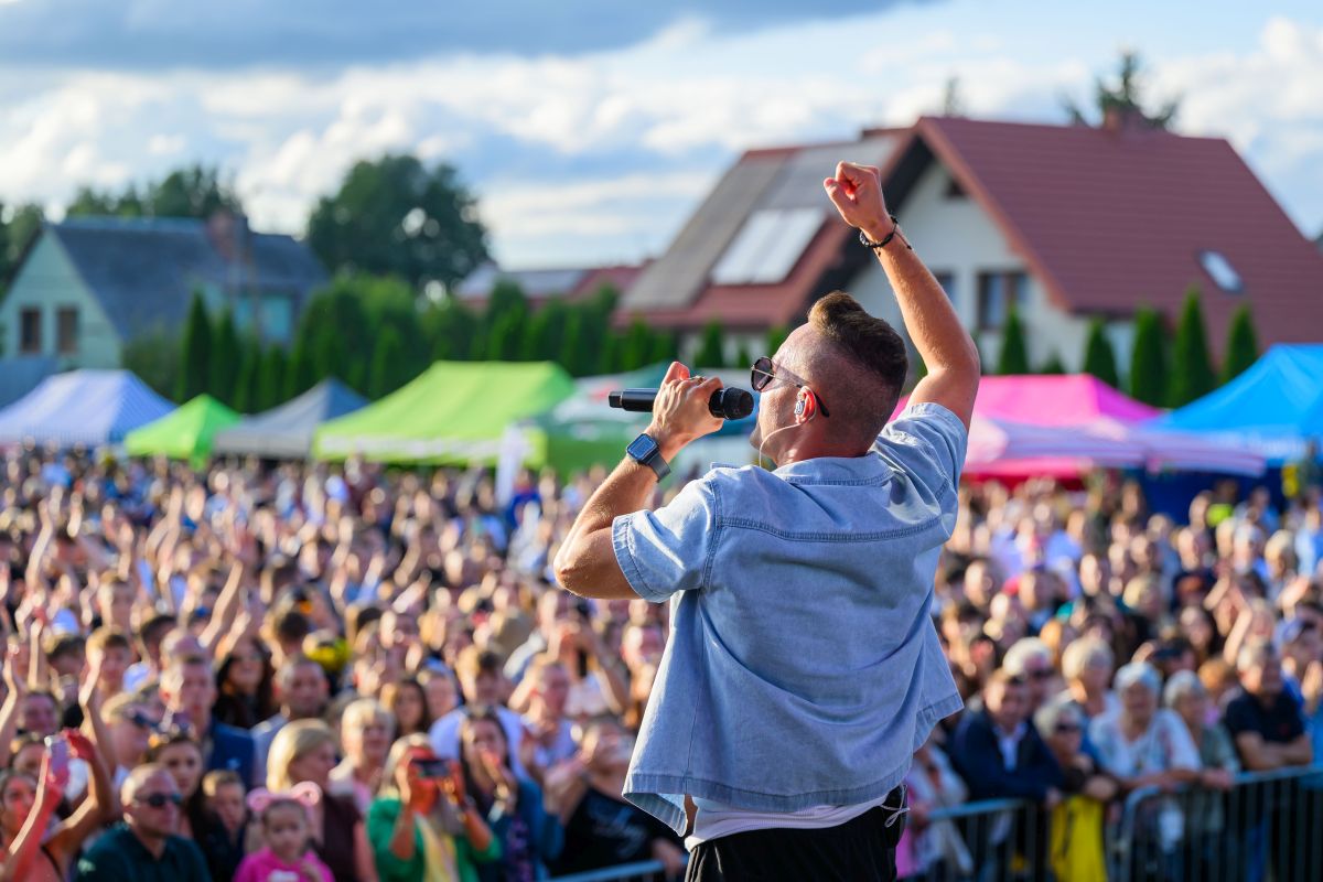 Mężczyzna śpiewa na scenie, trzyma mikrofon. Tłum ludzi, kolorowe namioty w tle. Jasny dzień, niebieskie niebo.