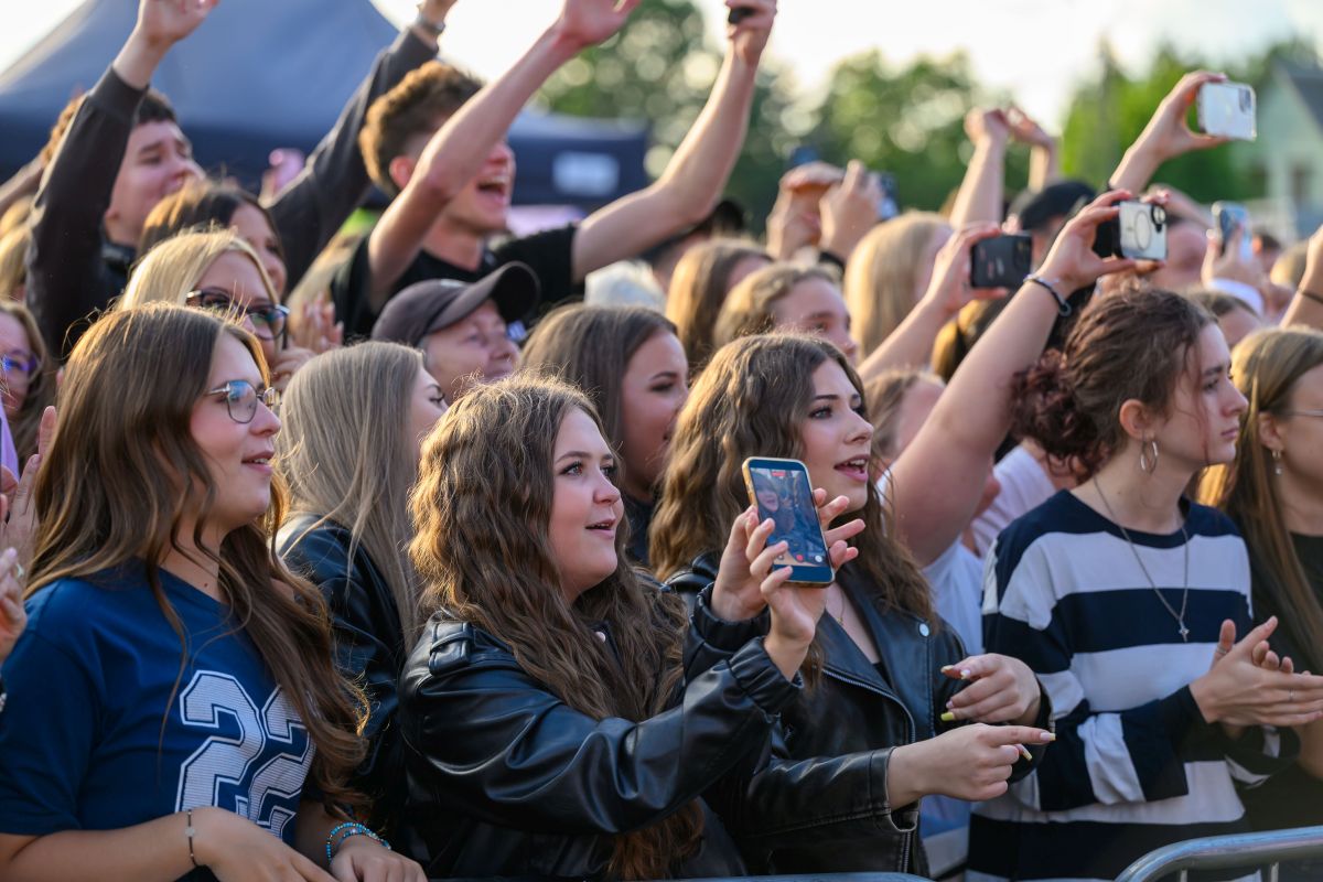 Grupa ludzi na koncercie, wznoszą ręce i robią zdjęcia. W tle zielone drzewa. Wiele osób ubranych w ciemne kolory.