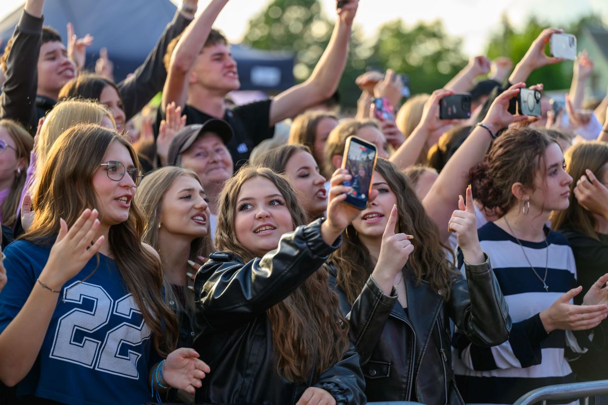 Grupa młodych ludzi z telefonami na koncercie, w radosnym nastroju. Kolorowe ubrania, oprawa w tle z zielenią. Tłum unosi ręce.