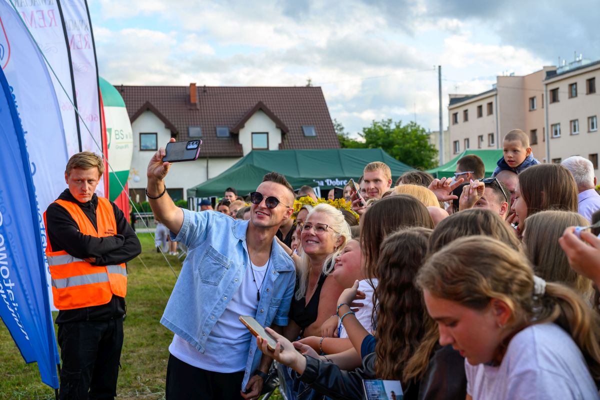 Mężczyzna robi selfie z grupą ludzi na festynie. W tle namioty i budynki, pochmurne niebo. Uczestnicy są uśmiechnięci i radośni.