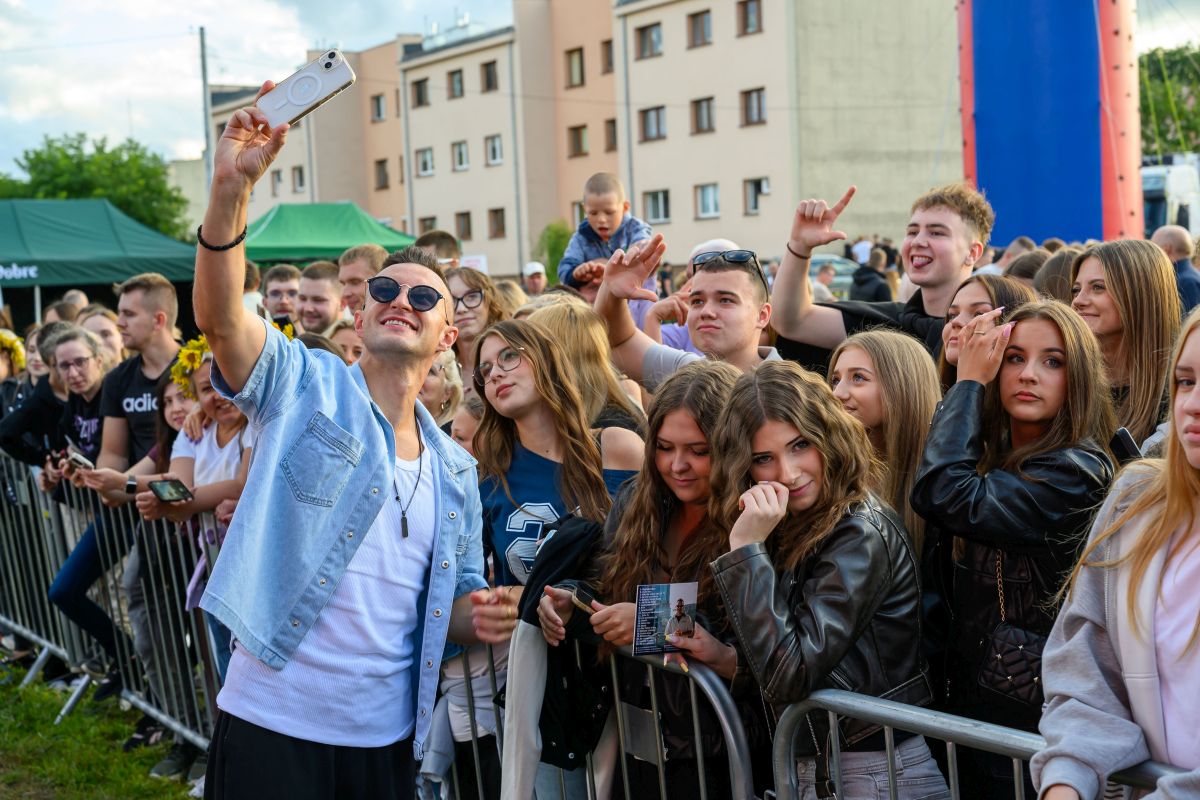 Mężczyzna w okularach robi selfie z grupą młodych ludzi za barierką. Uśmiechnięte twarze, różnorodne stroje, kolorowe tło. W tle budynki i namioty.
