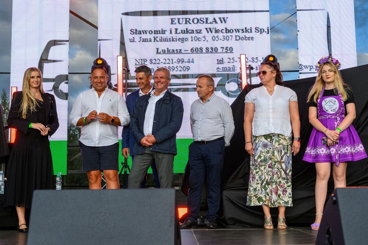 Grupa ludzi na scenie ubrana w casualowe i tradycyjne stroje. Tło to jasny ekran z tekstem. Pogoda jest pochmurna.