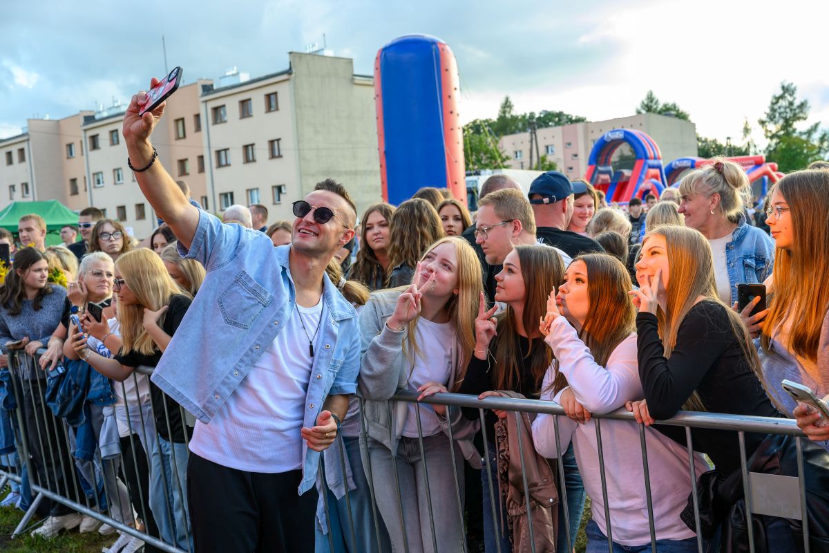 Młody mężczyzna w okularach przeciwsłonecznych robi selfie z grupą nastolatków. W tle kolorowe budynki i dmuchawce. Tłum ludzi uśmiecha się i pozuje.