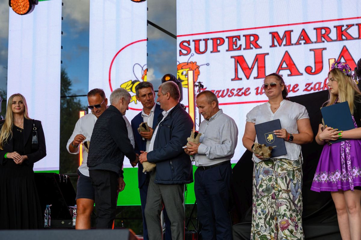 Grupa osób na scenie podczas ceremonii wręczenia nagród. Tło to ekran z logo SUPER MARKET MAJA. Osoby ubrani są elegancko, otoczeni są kolorowym oświetleniem.