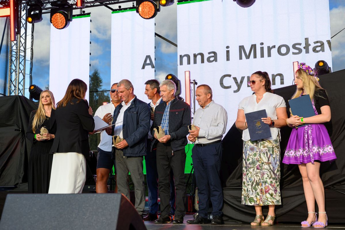 Grupa ludzi stoi na scenie, odbierając nagrody. Tło to duży ekran LED z jasnym niebem. Na zdjęciu dominują kolory: biały, czarny i fioletowy.