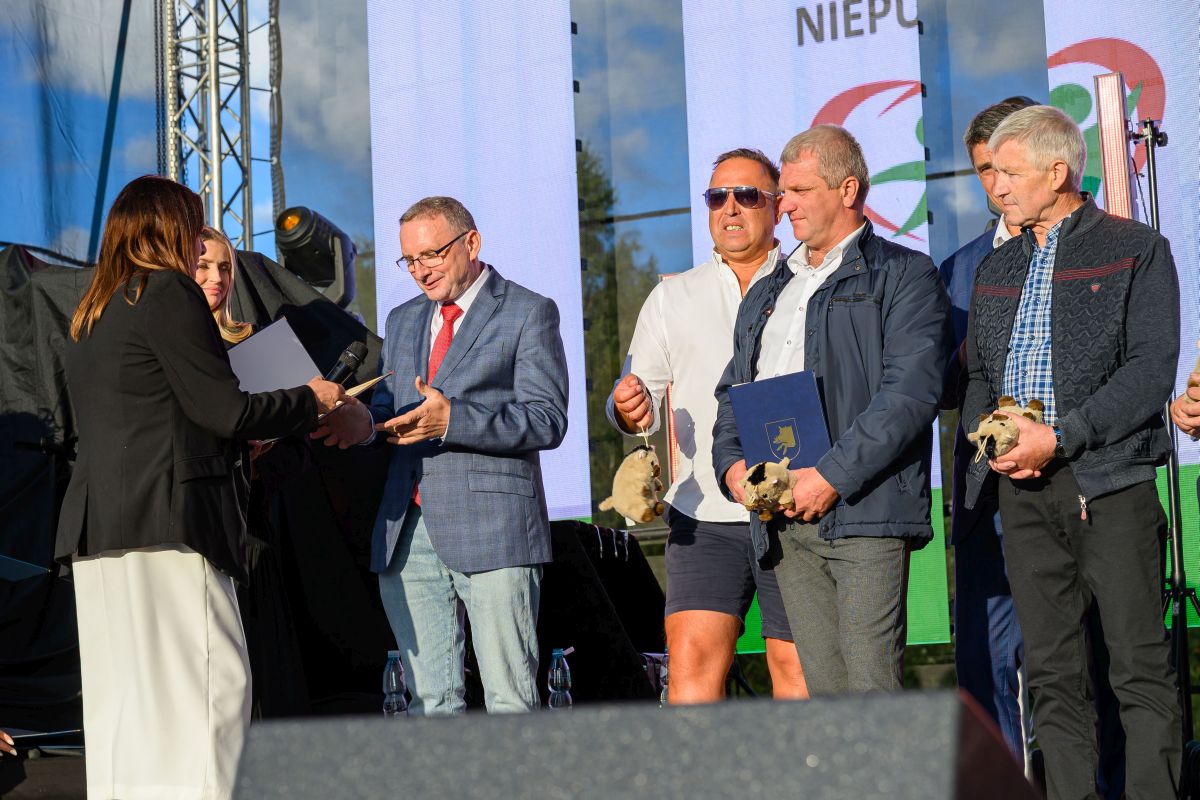 Na scenie grupa osób odbiera nagrody od kobiety w ciemnym stroju. W tle widoczny jest duży ekran i sprzęt oświetleniowy. Dominuje jasne oświetlenie i oficjalny charakter wydarzenia.