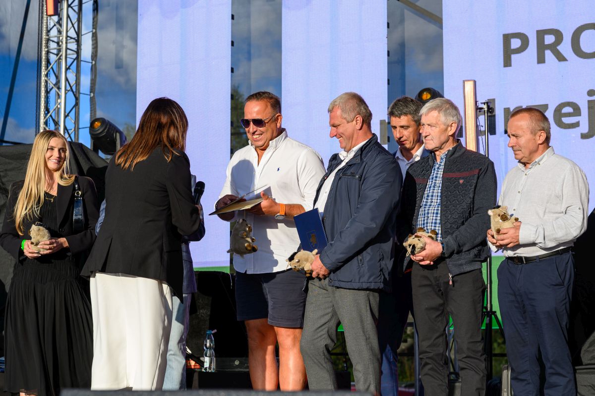 Grupa osób stoi na scenie, trzymając trofea. Tło to duży ekran, który wyświetla tekst. Atmosfera jest formalna, ale pogodna.