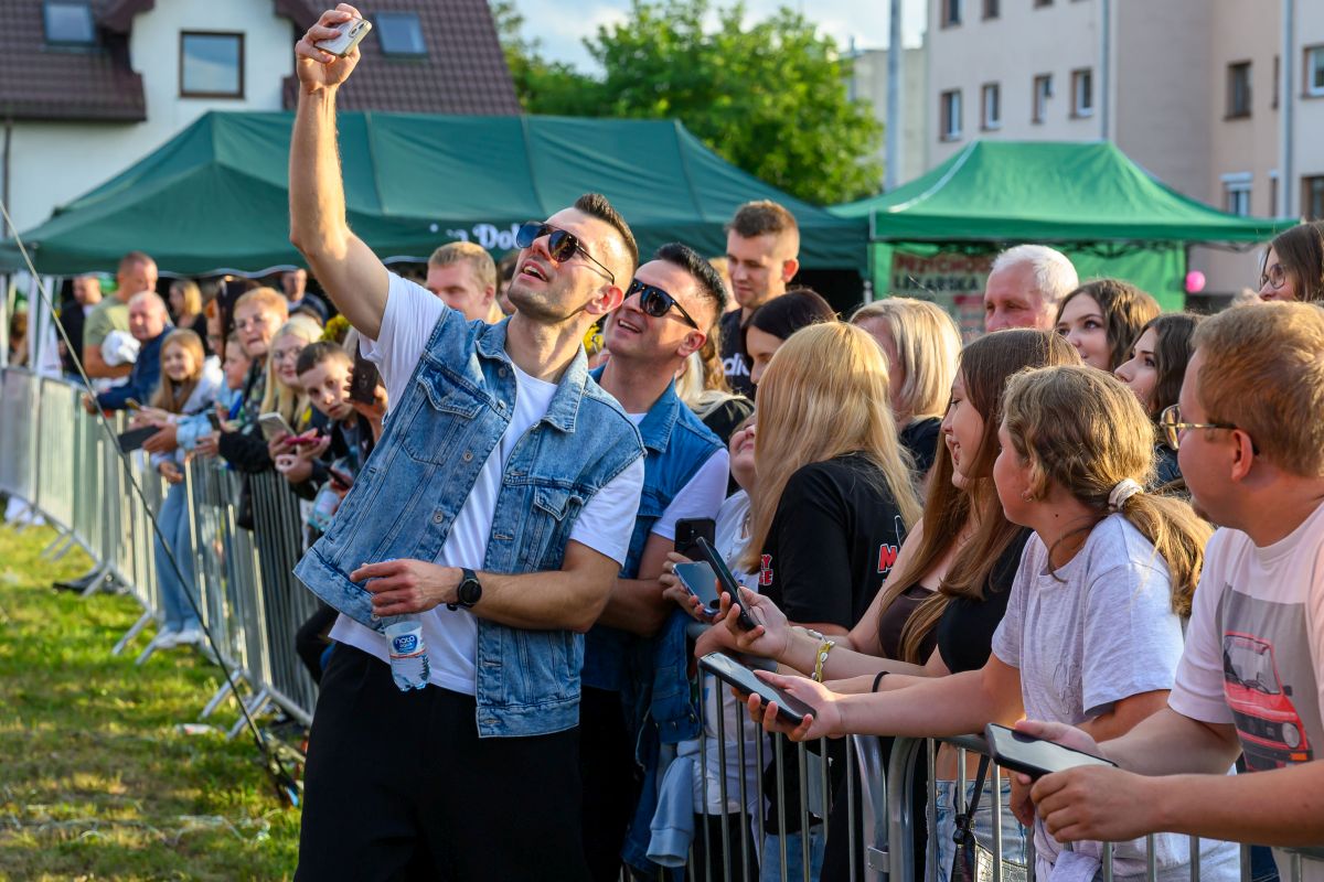 Na zdjęciu grupa osób robi selfie na tle zielonych namiotów. Ludzie stoją za barierkami, widać także budynki w tle. Dominuje kolor zielony i różnorodność ubrań uczestników.