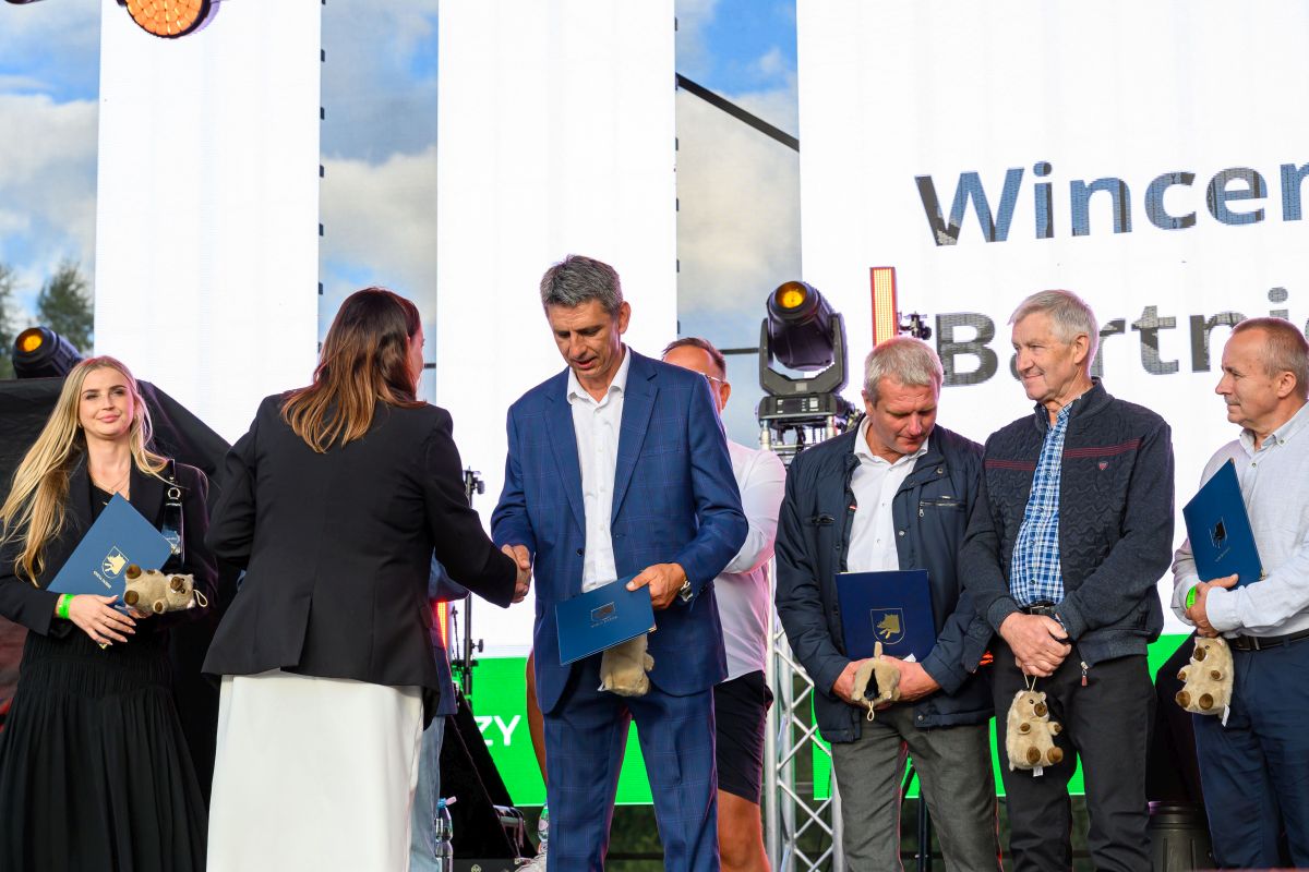 Grupa ludzi stoi na scenie podczas ceremonii wręczenia nagród. Tło z jasnym niebem i białym banerem. Osoby trzymają niebieskie teczki i maskotki.